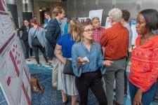 A young woman presents a trifold board research project to several onlookers.